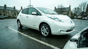 A second-hand electric car listing on a smartphone screen beside a charging cable plugged into a white Nissan Leaf